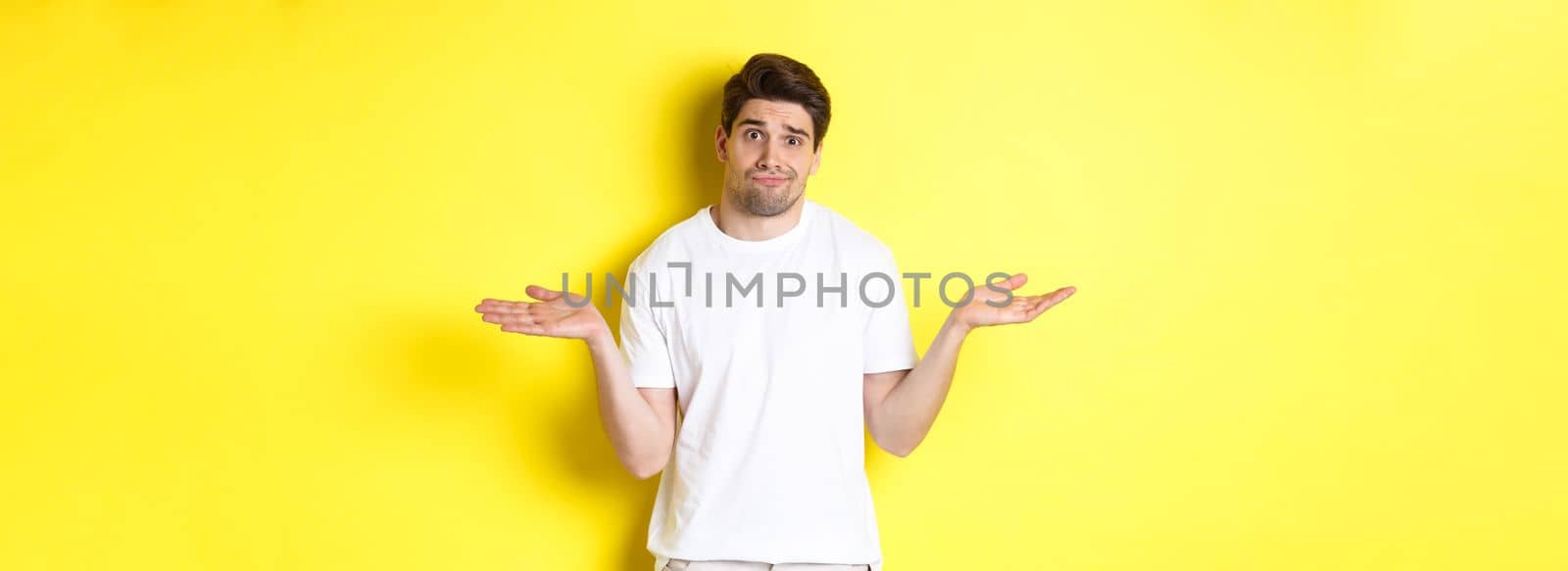 Clueless man in white t-shirt, shrugging and looking puzzled, dont know anything, standing over yellow background.