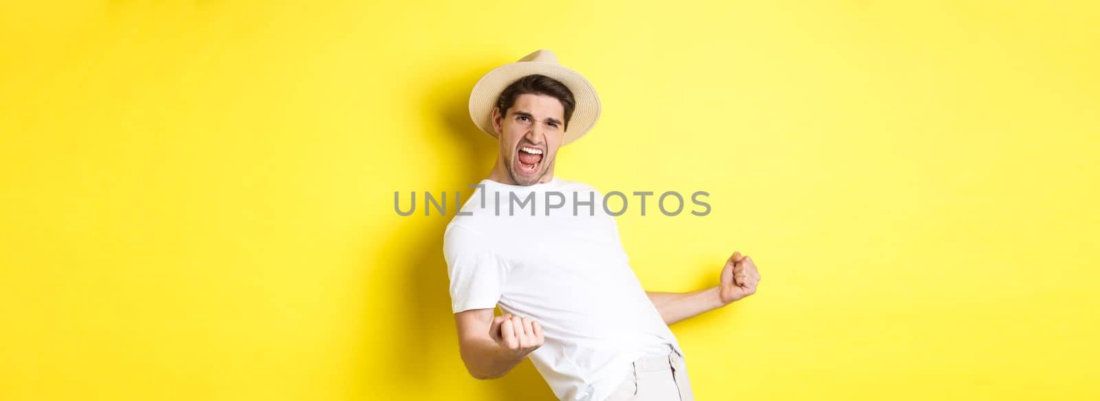Concept of tourism and vacation. Satisfied young male tourist celebrating, winning something and rejoicing, making fist pump and shouting yes, standing against yellow background.