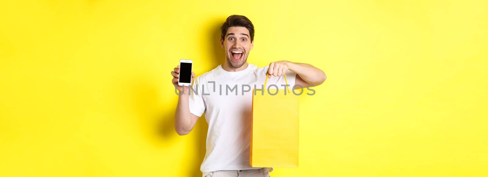 Concept of mobile banking and cashback. Young happy guy holding shopping bag and showing smartphone screen, yellow background by Benzoix