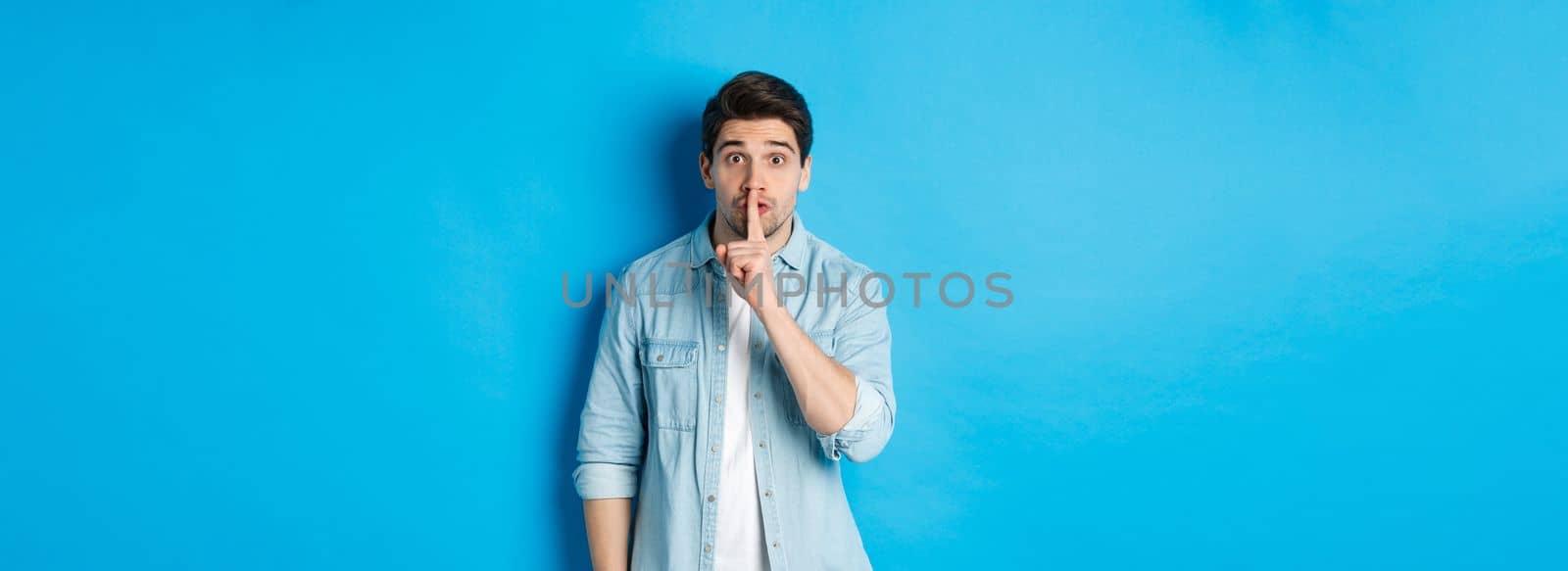 Portrait of excited man asking to keep quiet, showing hush taboo sign and looking nervously at camera, standing against blue background.