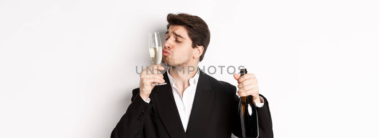 Close-up of handsome man in suit, kissing glass with champagne, getting drunk on a party, standing against white background by Benzoix