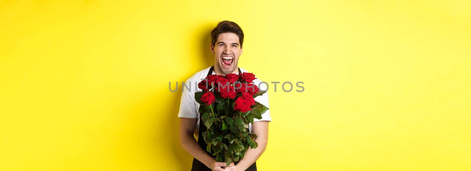 Funny young salesman in black apron holding bouquet of roses, florist laughing and standing over yellow background by Benzoix