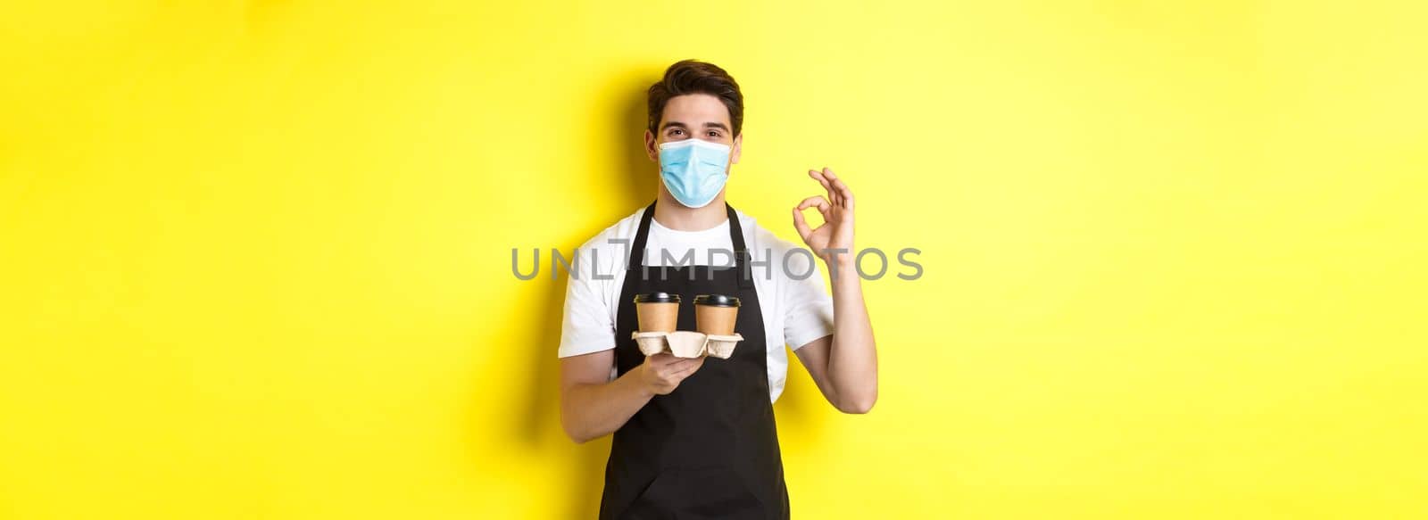 Concept of covid-19, cafe and social distancing. Barista in medical mask and black apron guarantee safety, holding takeaway cups of coffee and showing OK sign, yellow background.