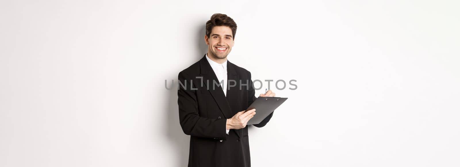 Portrait of confident businessman in black suit, signing documents and smiling, standing happy against white background.