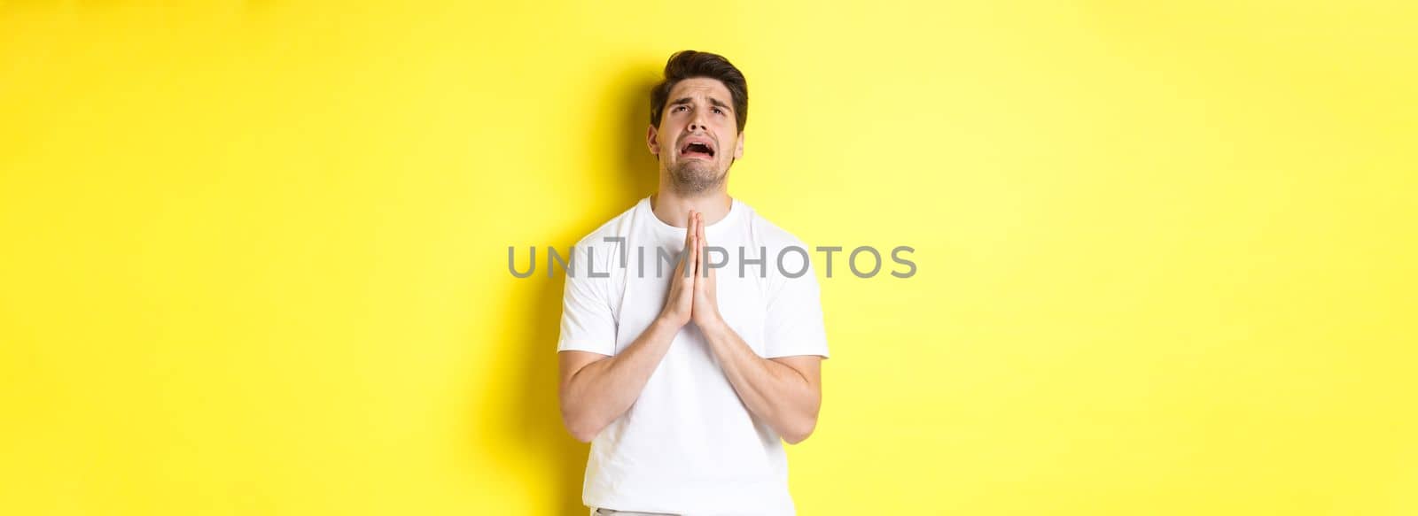 Desperate man pleading God, holding hands in pray and looking up distressed, standing over yellow background. Copy space