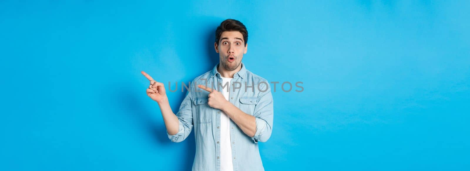 Portrait of surprised bearded guy in casual outfit pointing fingers right, showing amazing promo offer, standing over blue background.