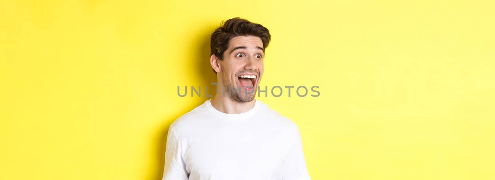 Image of happy man checking out promo, looking left with amazement, standing in white t-shirt against yellow background by Benzoix