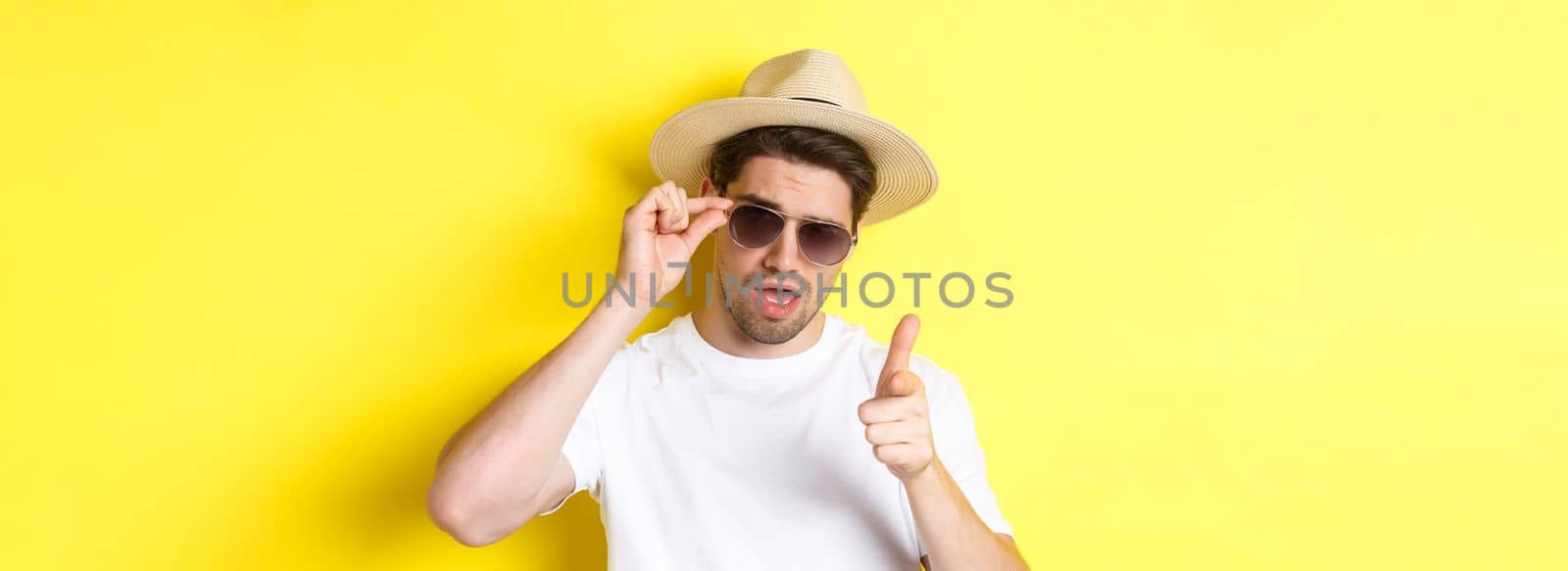 Concept of tourism and vacation. Cool and sassy man flirting with you, wearing sunglasses and pointing finger at camera, standing over yellow background by Benzoix