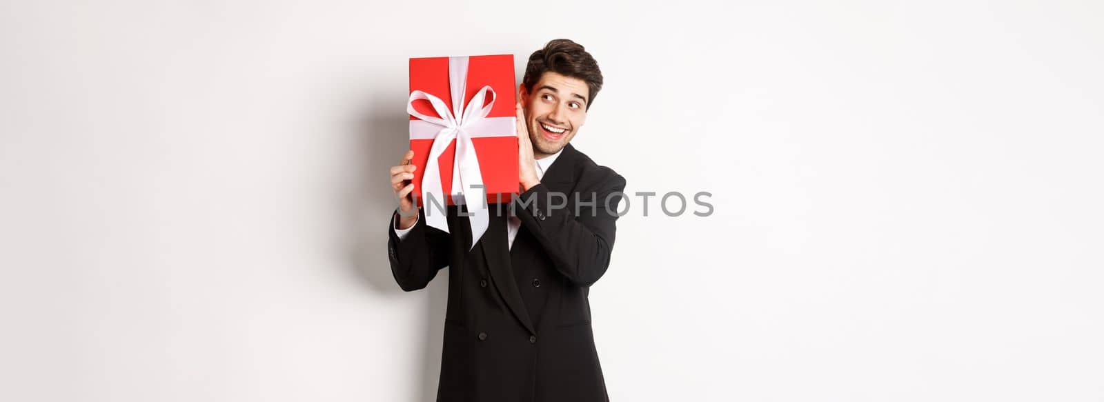 Concept of christmas holidays, celebration and lifestyle. Image of excited man enjoying new year, shaking gift box to guess what inside, standing against white background.