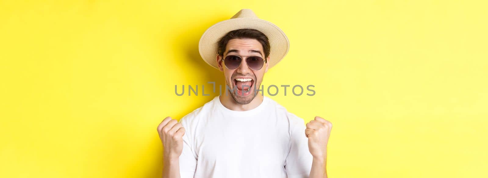 Concept of tourism and holidays. Lucky man tourist winning tickets, making fist pump and saying yes, wearing vacation clothes, standing over yellow background by Benzoix