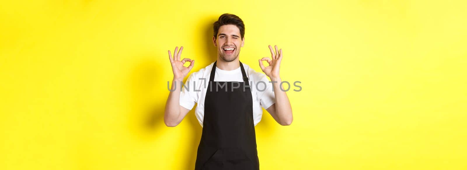 Young male barista in black apron showing OK signs and smiling, guarantee quality in coffee shop, standing over yellow background.