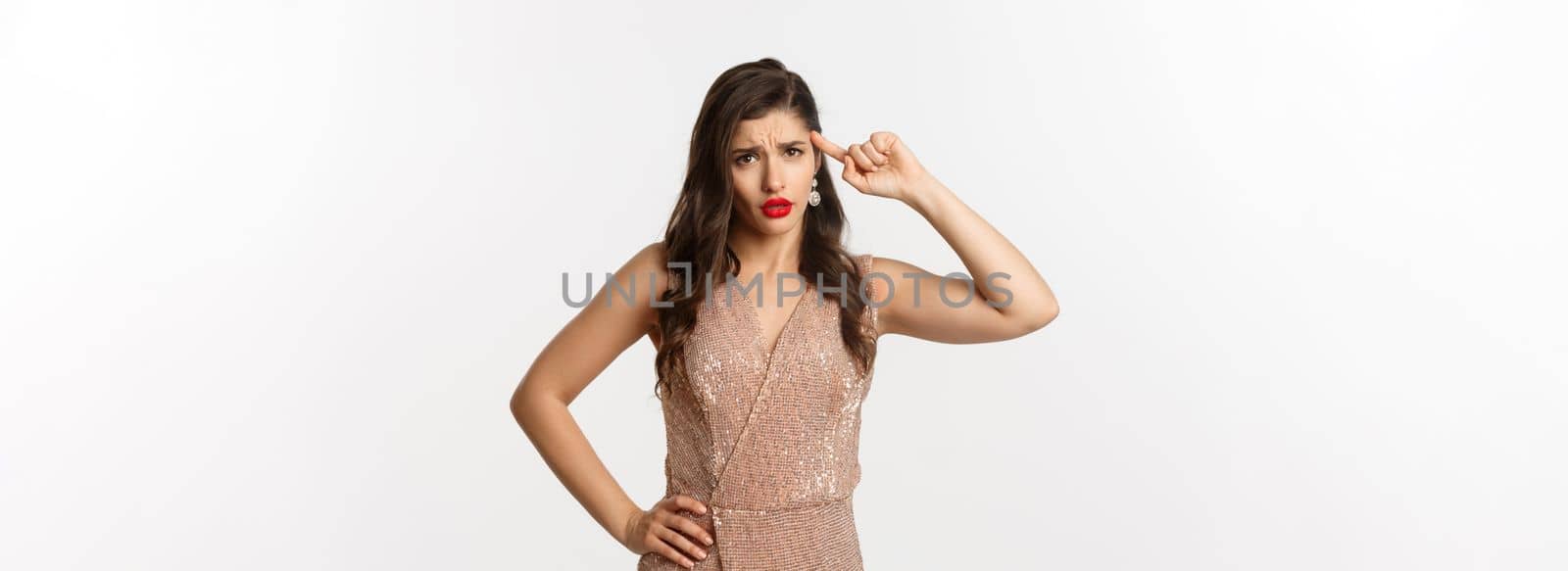 Concept of casino, celebration and party. Woman looking with disdain and roll finger on head, scolding someone, are you stupid gesture, standing in dress over white background.