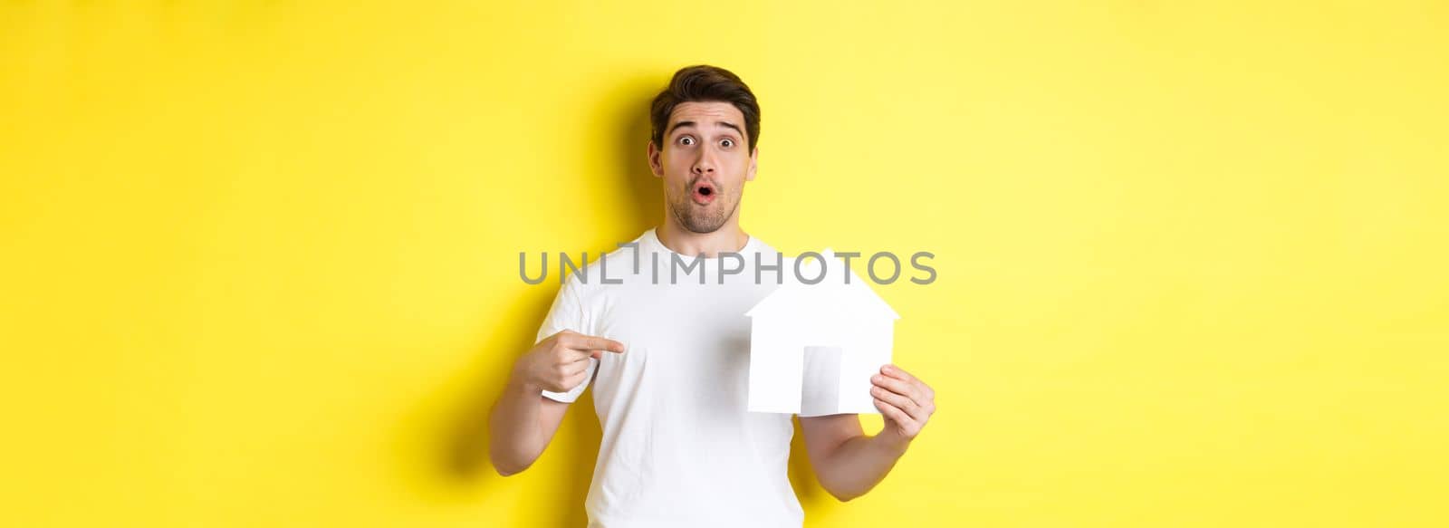 Real estate concept. Excited man pointing finger at paper house model, searching for flat, standing over yellow background.