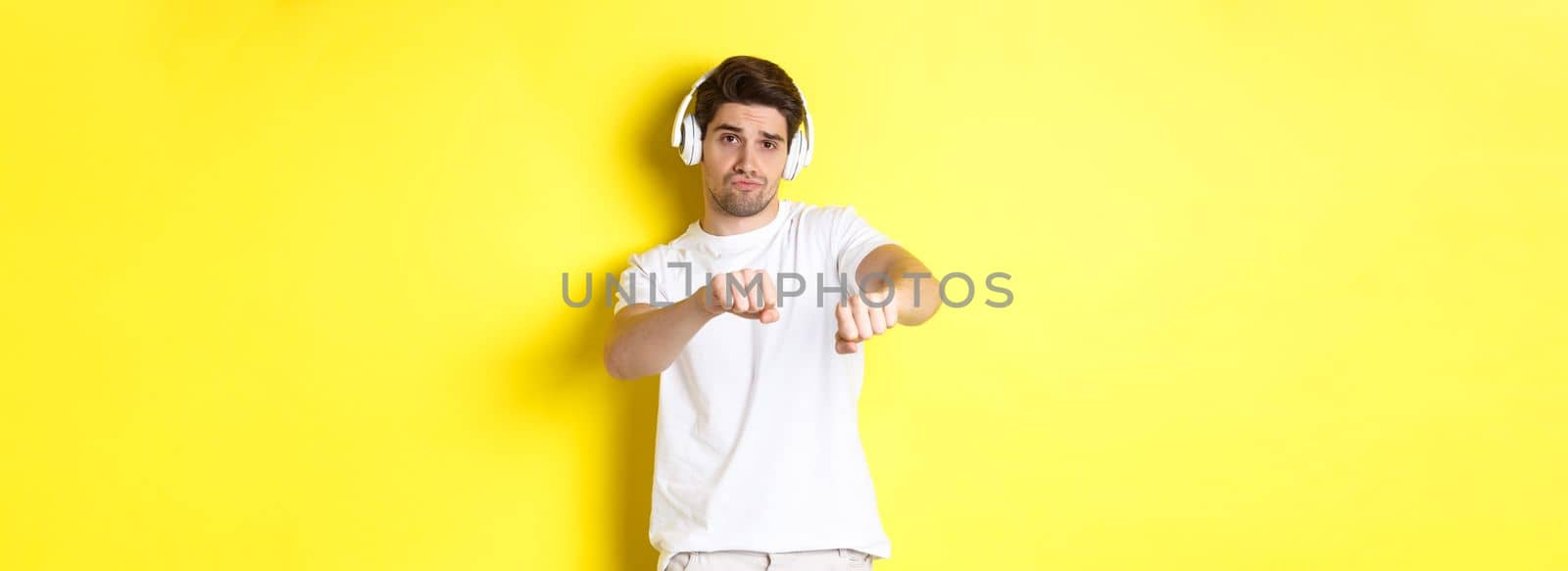 Cool guy listening music in headphones and dancing, standing in white clothes against yellow studio background by Benzoix