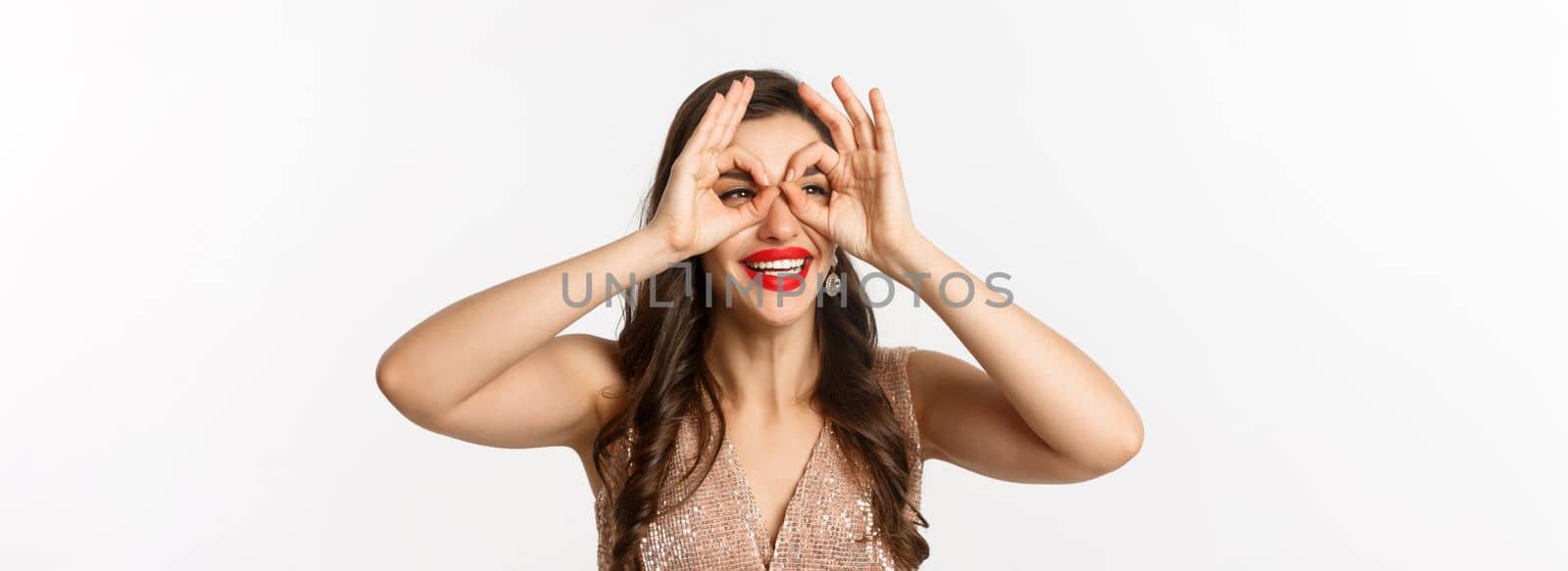 Concept of New Year celebration and winter holidays. Close-up of beautiful brunette woman in dress, red lips, making hand binoculars and staring left, white background.