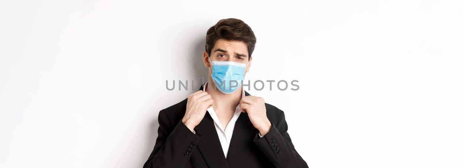 Concept of covid-19, business and social distancing. Image of confident handsome man in trendy suit and medical mask, looking sassy, standing against white background by Benzoix