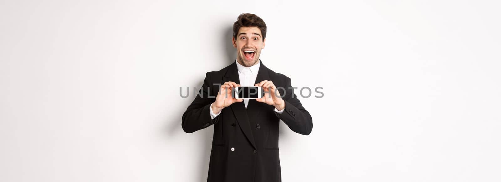 Image of cheerful, handsome man in black suit, showing smarthone screen and looking amazed, standing against white background.