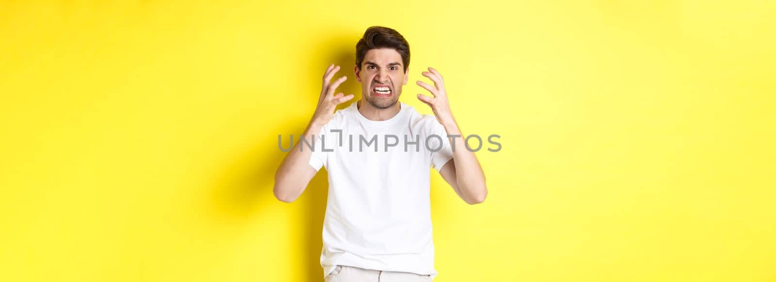 Angry man looking mad, grimacing and shaking hands furious, standing outraged against yellow background.