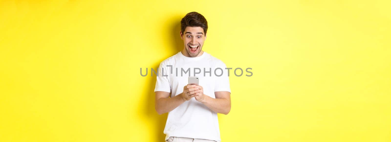Surprised man reading text message on mobile phone, looking amazed and happy at smartphone screen, standing over yellow background by Benzoix
