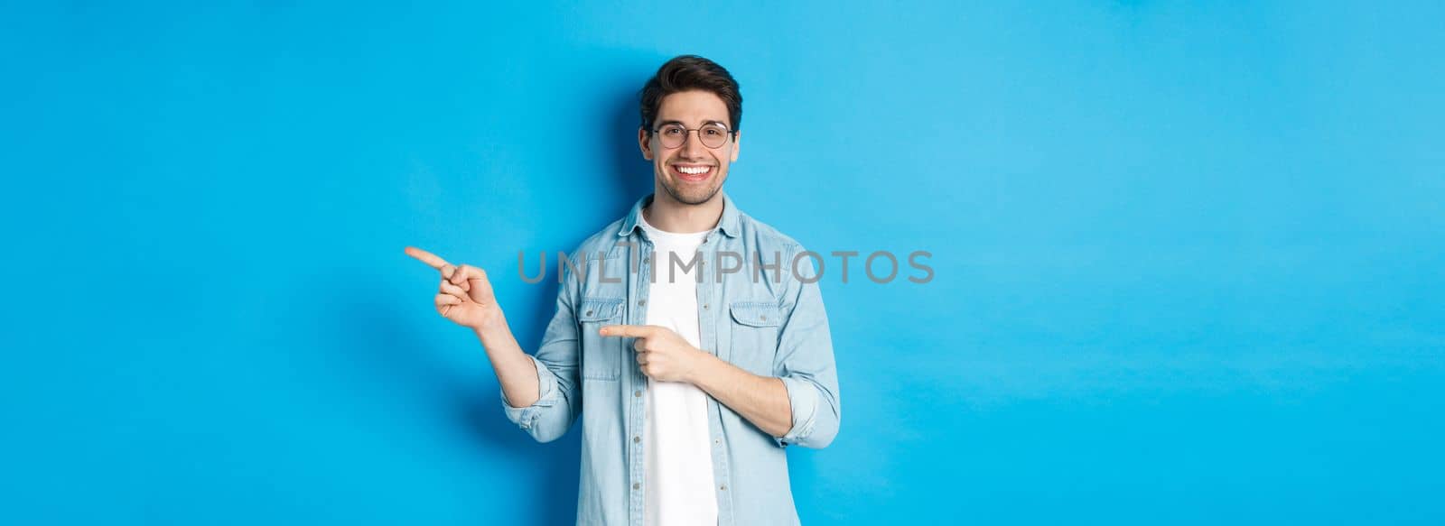 Handsome caucasian male model in glasses, pointing fingers right at your logo, showing copy space, standing over blue background by Benzoix