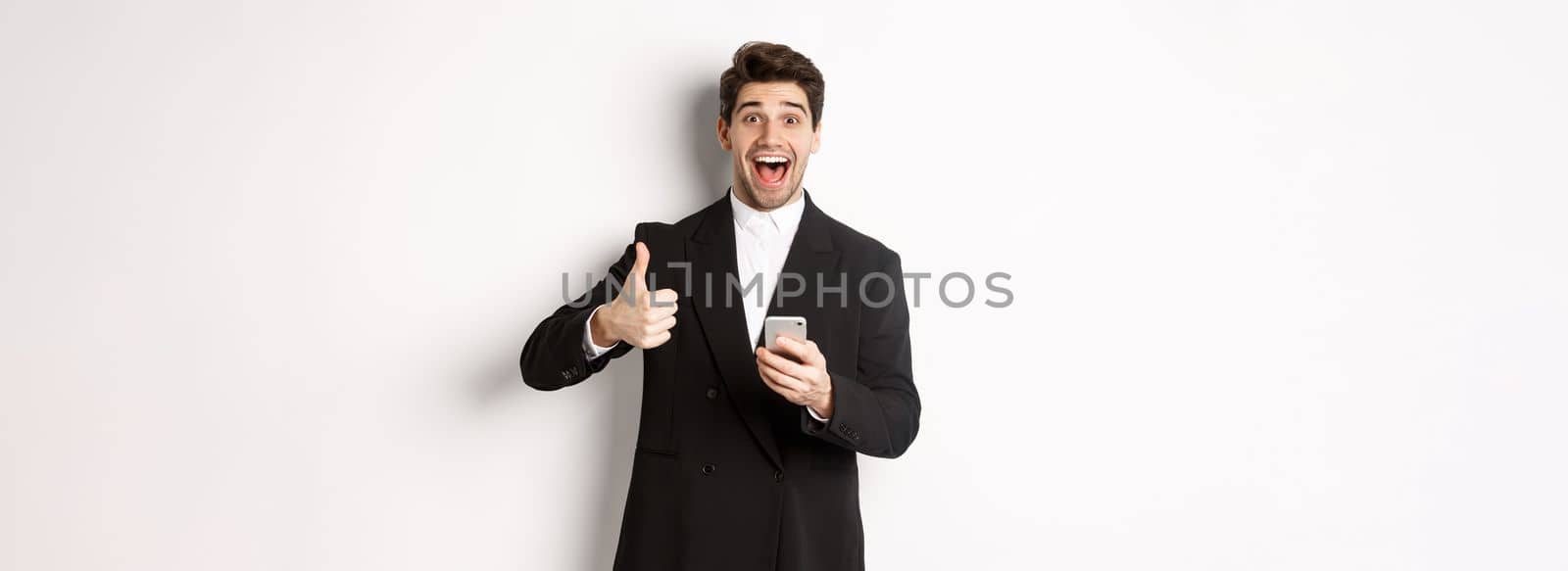 Portrait of handsome man in trendy suit showing thumbs-up in approval, using mobile phone app, smiling pleased, standing over white background.