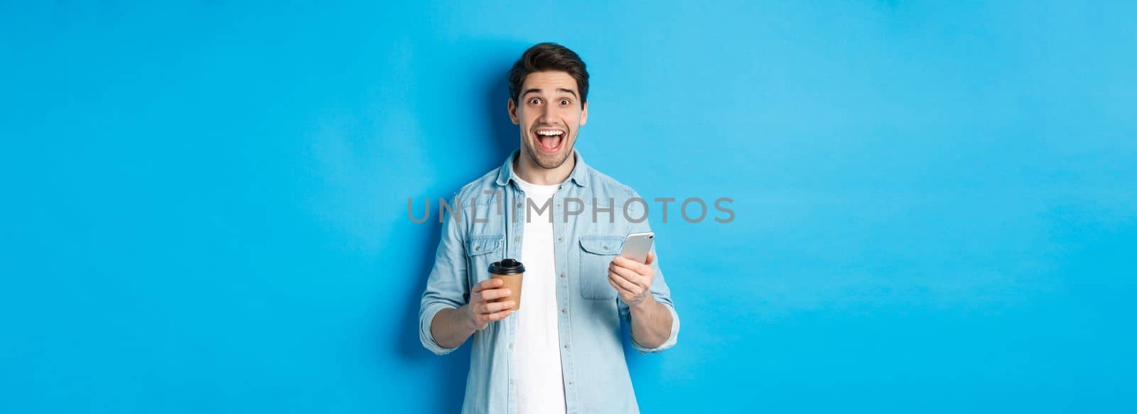 Happy young man drinking coffee and using mobile phone, looking excited, standing against blue background by Benzoix