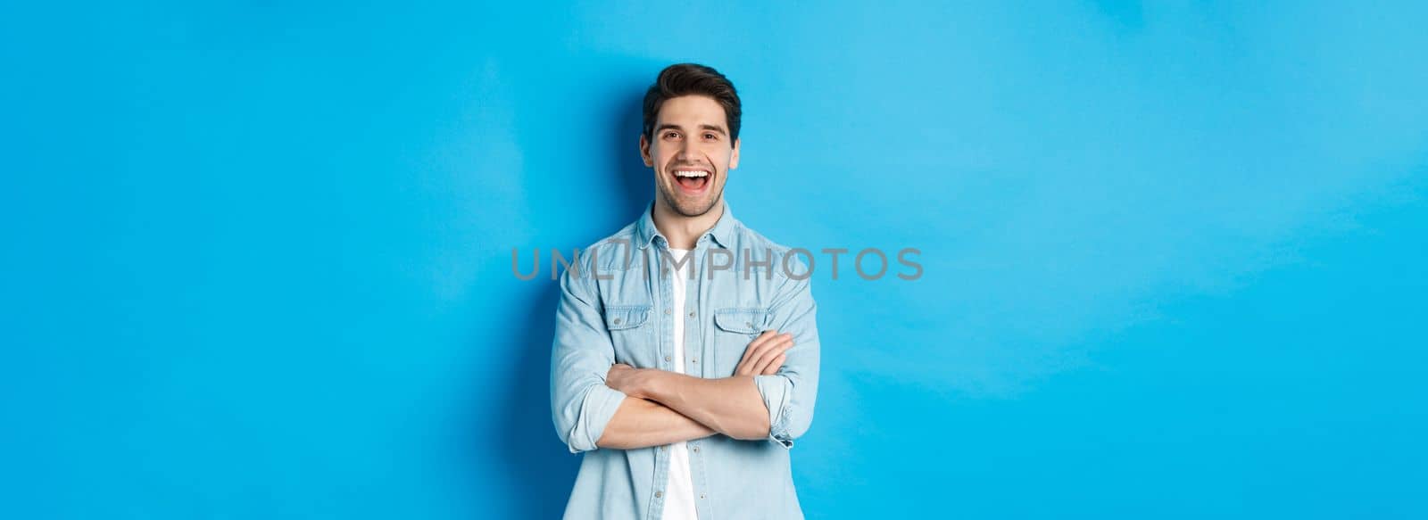 Handsome guy with beard, wearing casual outfit, laughing and looking at something funny, standing against blue background.