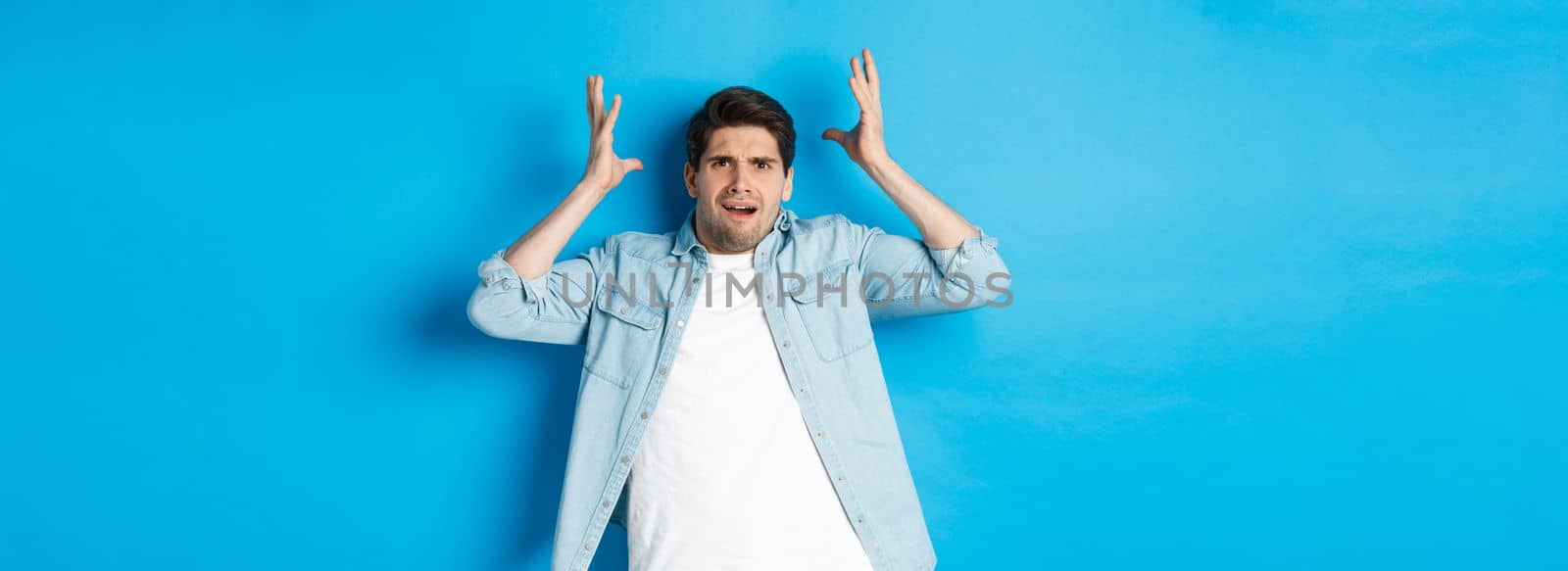 Confused and bothered man holding hands near head, looking frustrated, standing upset against blue background.