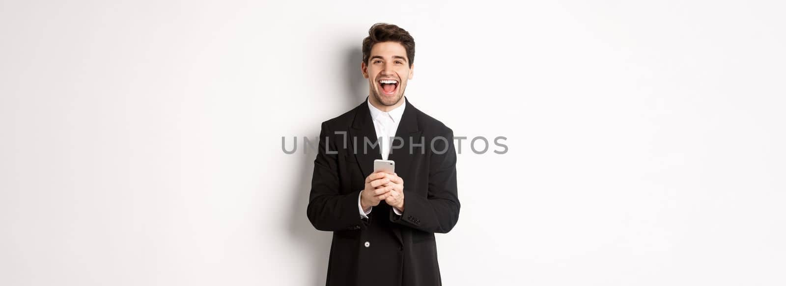 Portrait of happy good-looking man, wearing black suit, laughing from happiness and using mobile phone, standing over white background by Benzoix