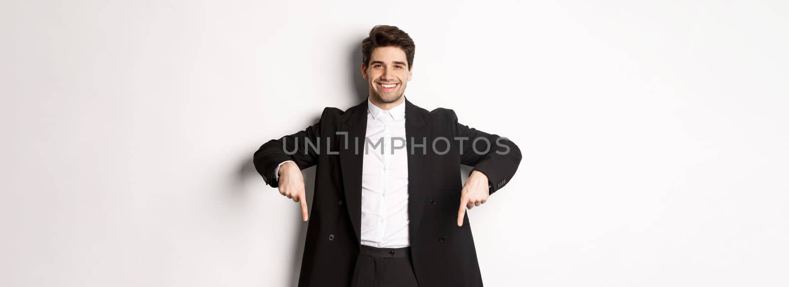 Portrait of good-looking stylish man in black suit, pointing fingers down and smiling, showing winter holidays promo, standing over white background by Benzoix
