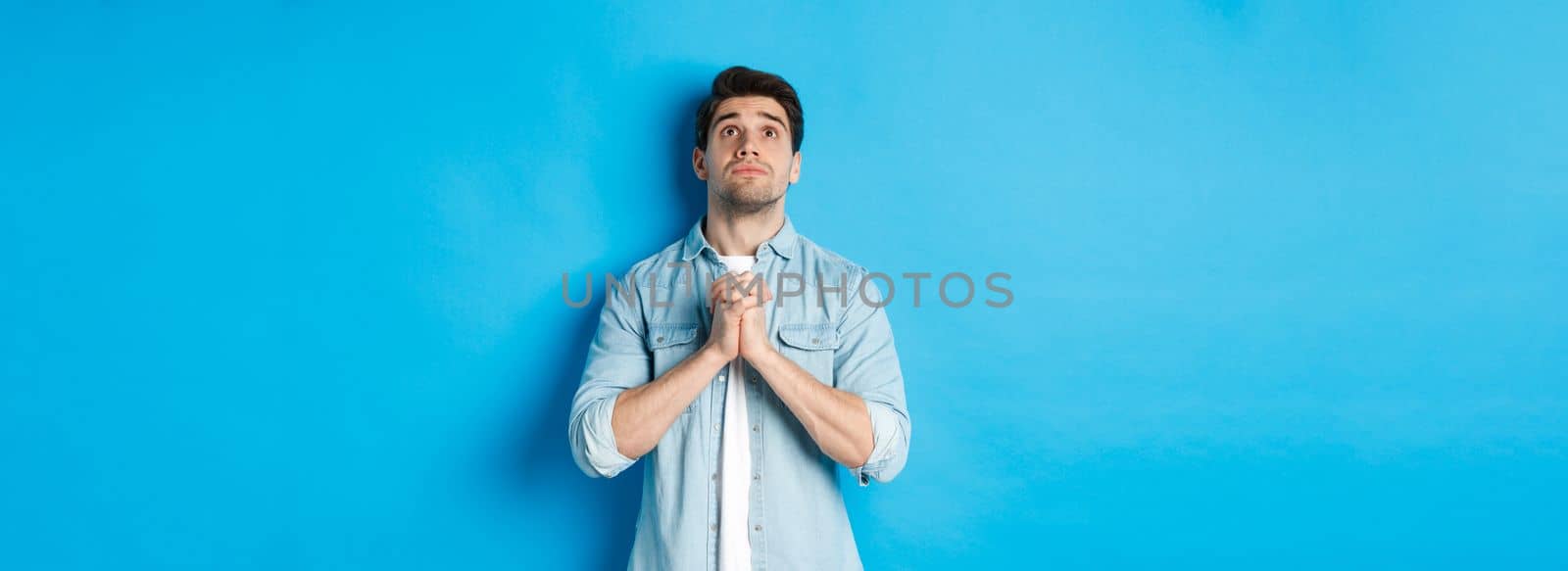 Hopeful young man looking up and praying God, begging for help, standing against blue background.