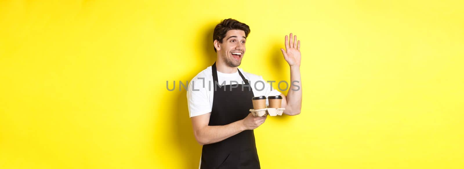 Friendly waiter in cafe waving hand at customer, holding takeaway coffee oder, standing against yellow background in black apron.