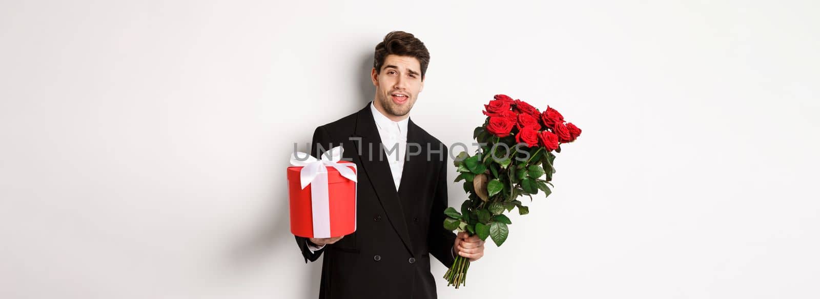 Concept of holidays, relationship and celebration. Handsome and confident man in black suit, going on a date, holding bouquet of roses and present, standing against white background by Benzoix