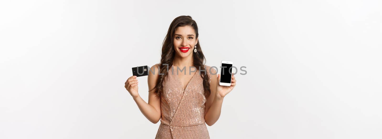 Online shopping and holidays concept. Attractive woman in party dress showing credit card and mobile screen, smiling happy, white background by Benzoix
