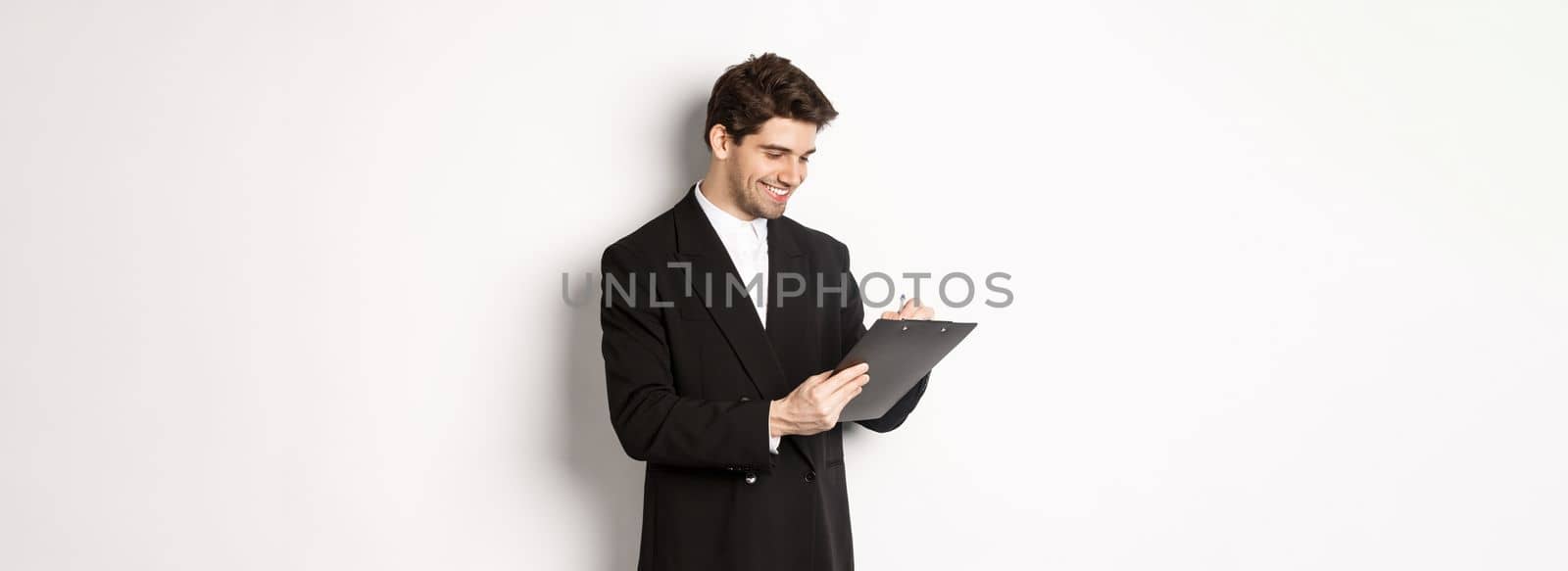 Image of handsome businessman in suit signing documents, looking at clipboard and smiling, standing against white background.