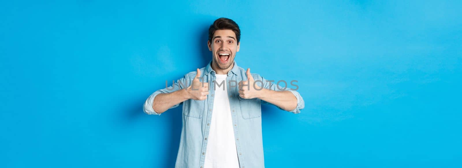 Smiling adult man showing thumbs up with excited face, like something awesome, approving product, standing against blue background by Benzoix