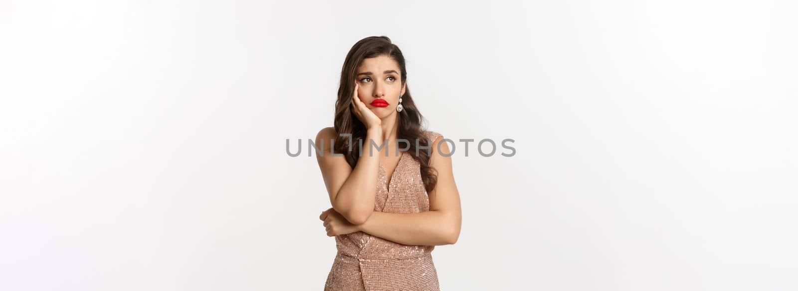 Bored and thoughtful young woman dreaming, looking away and imaging something with sad face, standing in glamour dress and waiting, standing over white background.
