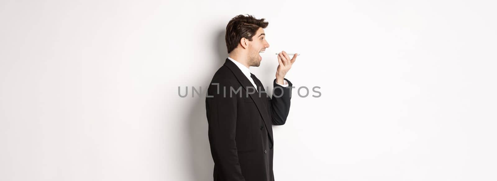 Profile shot of handsome businessman in black suit talking on speakerphone, smiling and looking happy, recording voice message, standing over white background.