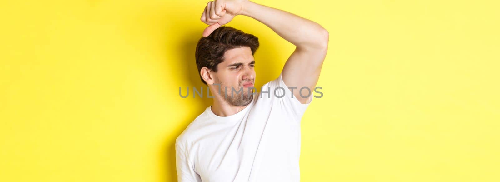 Man in sweat smelling his armpit, standing in white t-shirt and grimacing from stinky clothes.