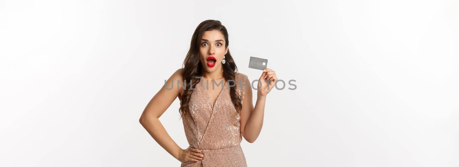 Holidays. christmas party and shopping concept. Amazed attractive woman with red lips, luxury dress, showing credit card and staring at camera, standing over white background by Benzoix