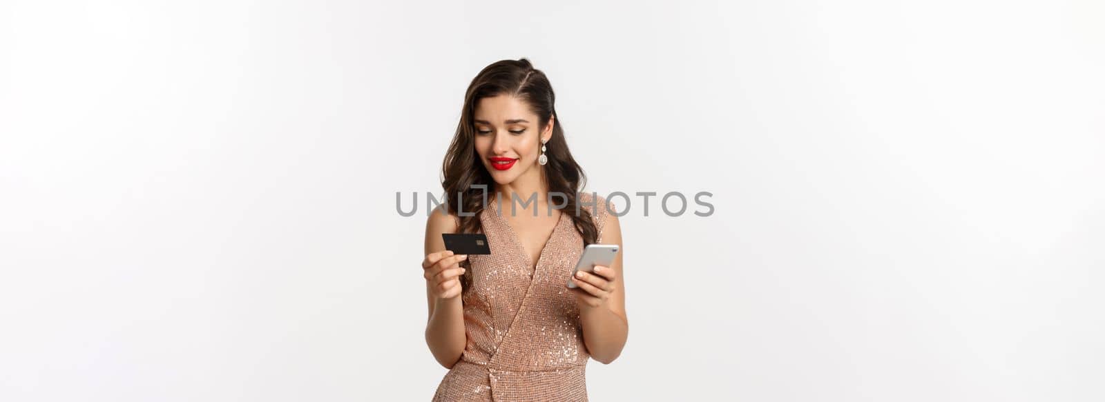 Online shopping and christmas concept. Elegant young woman paying for internet purchase with smartphone and credit card, standing in luxury dress over white background.