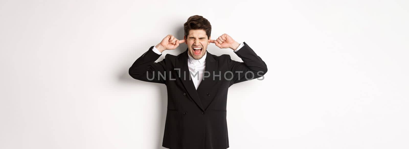 Portrait of annoyed and bothered businessman in black suit, shut ears and yelling, complaining loud noise, standing against white background.