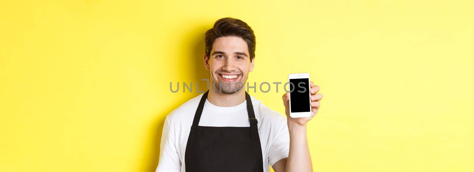 Close-up of handsome waiter in black apron showing smartphone screen, recommending app, standing over yellow background by Benzoix