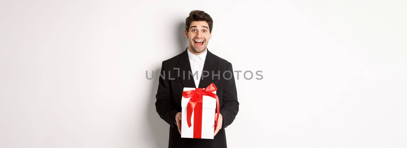 Concept of christmas holidays, celebration and lifestyle. Image of handsome guy in black suit looking excited, have a gift, standing against white background.