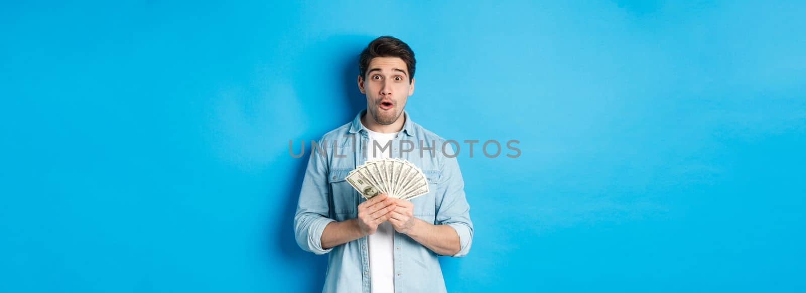 Concept of finance, credit and banking. Surprised man holding money, looking at camera wondered, standing over blue background.
