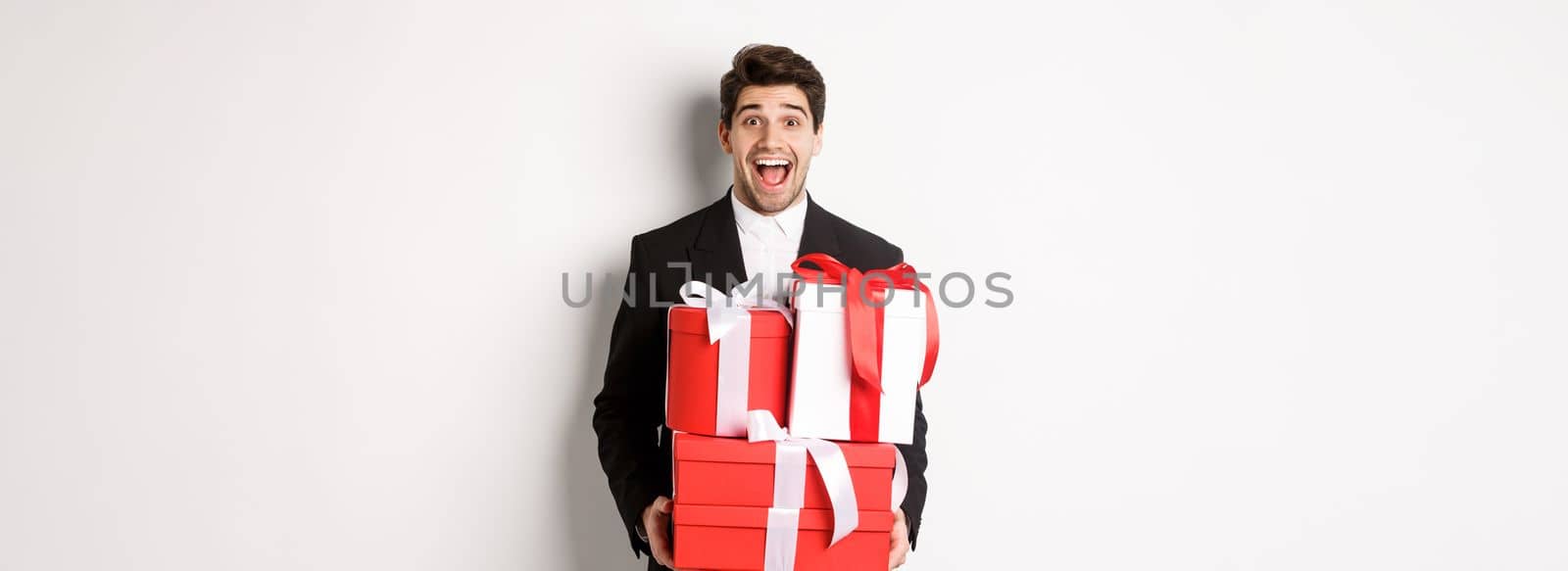 Concept of christmas holidays, celebration and lifestyle. Image of handsome amazed guy in suit, holding new year presents and smiling, standing against white background.