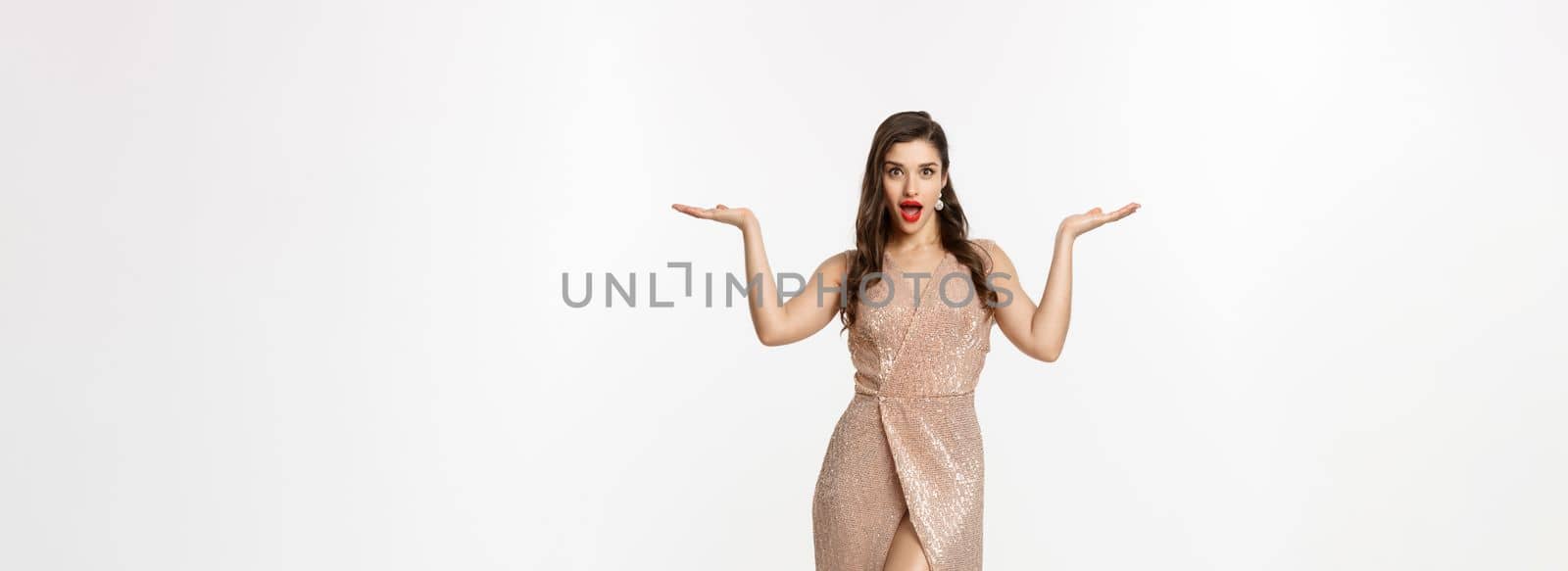 Christmas party and celebration concept. Attractive brunette female model in elegant dress, showing two offers, raising hands and holding something on copy space, white background by Benzoix