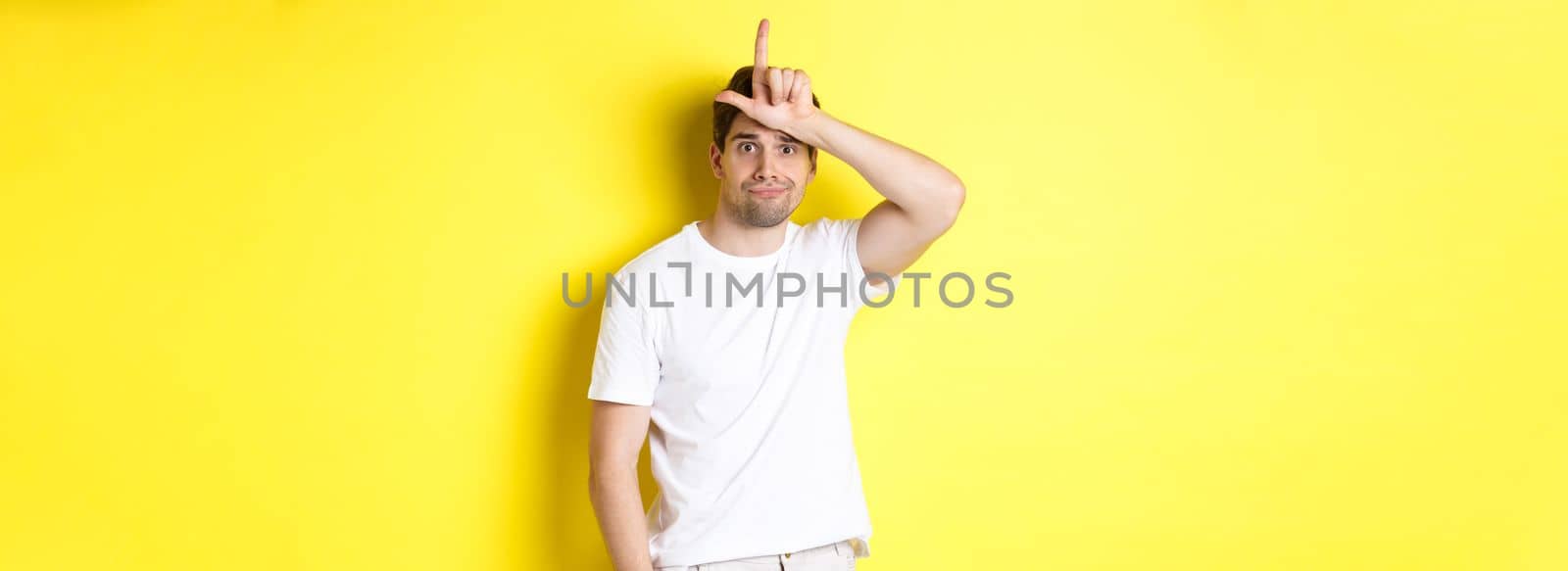 Awkward guy showing loser sign on forehead, looking sad and gloomy, standing in white t-shirt against yellow background.