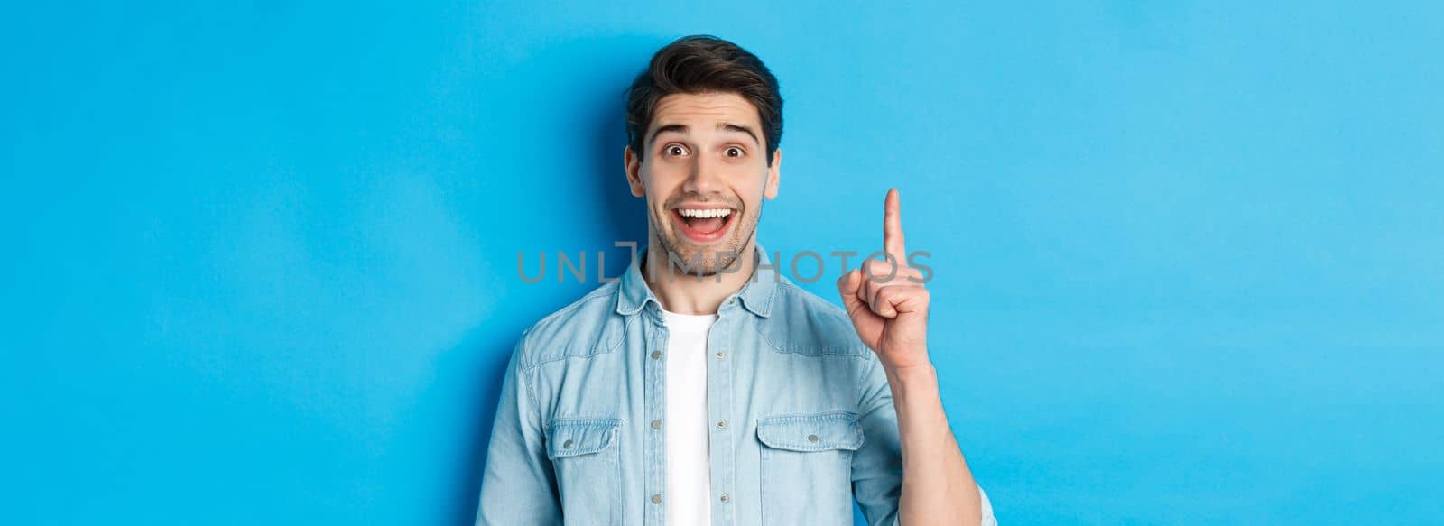 Close-up of handsome bearded guy smiling, showing finger number one, standing over blue background.