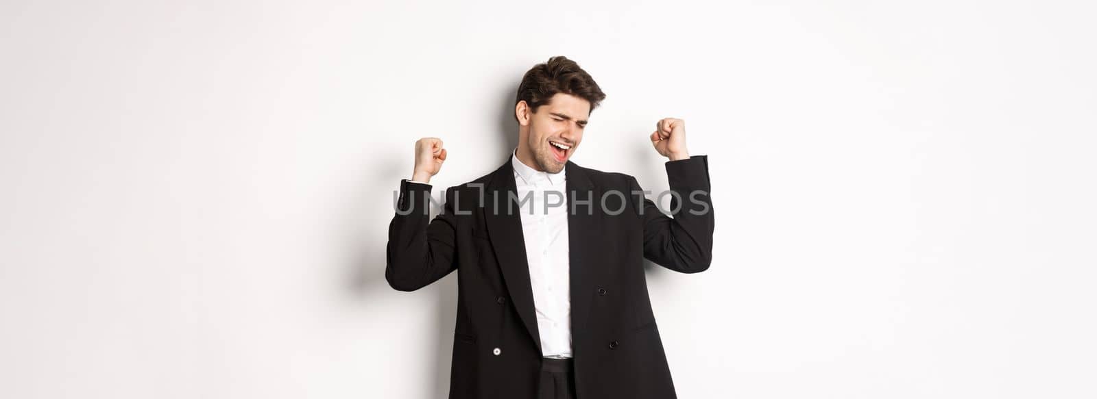 Image of successful and pleased handsome man in suit, rejoicing and making fist pump signs, dancing from happiness, standing over white background by Benzoix